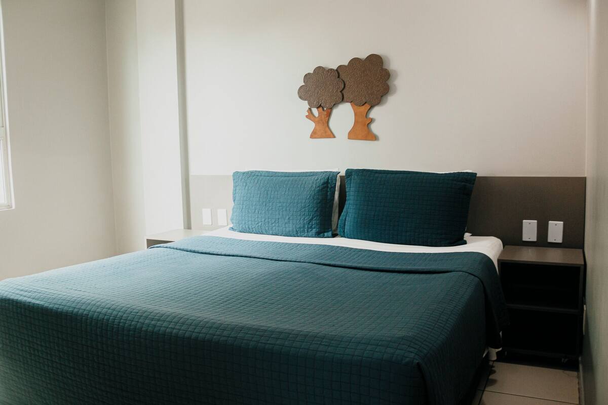 A neatly made bed with a teal bedspread and pillows, a wall-mounted tree decoration, and a nightstand with two white switches.