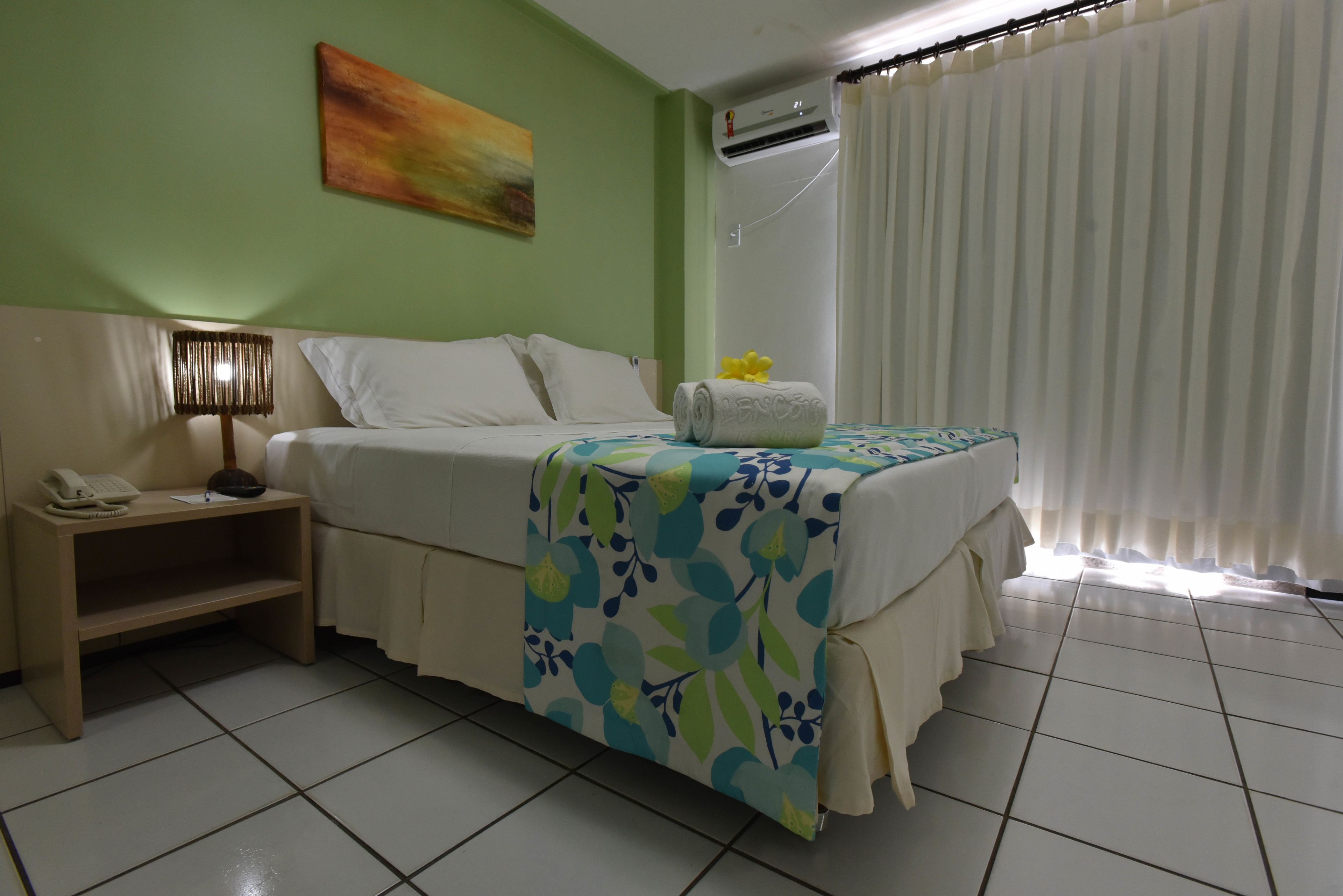A hotel room with a bed, a nightstand with a lamp, a painting on the wall, and an air conditioning unit.