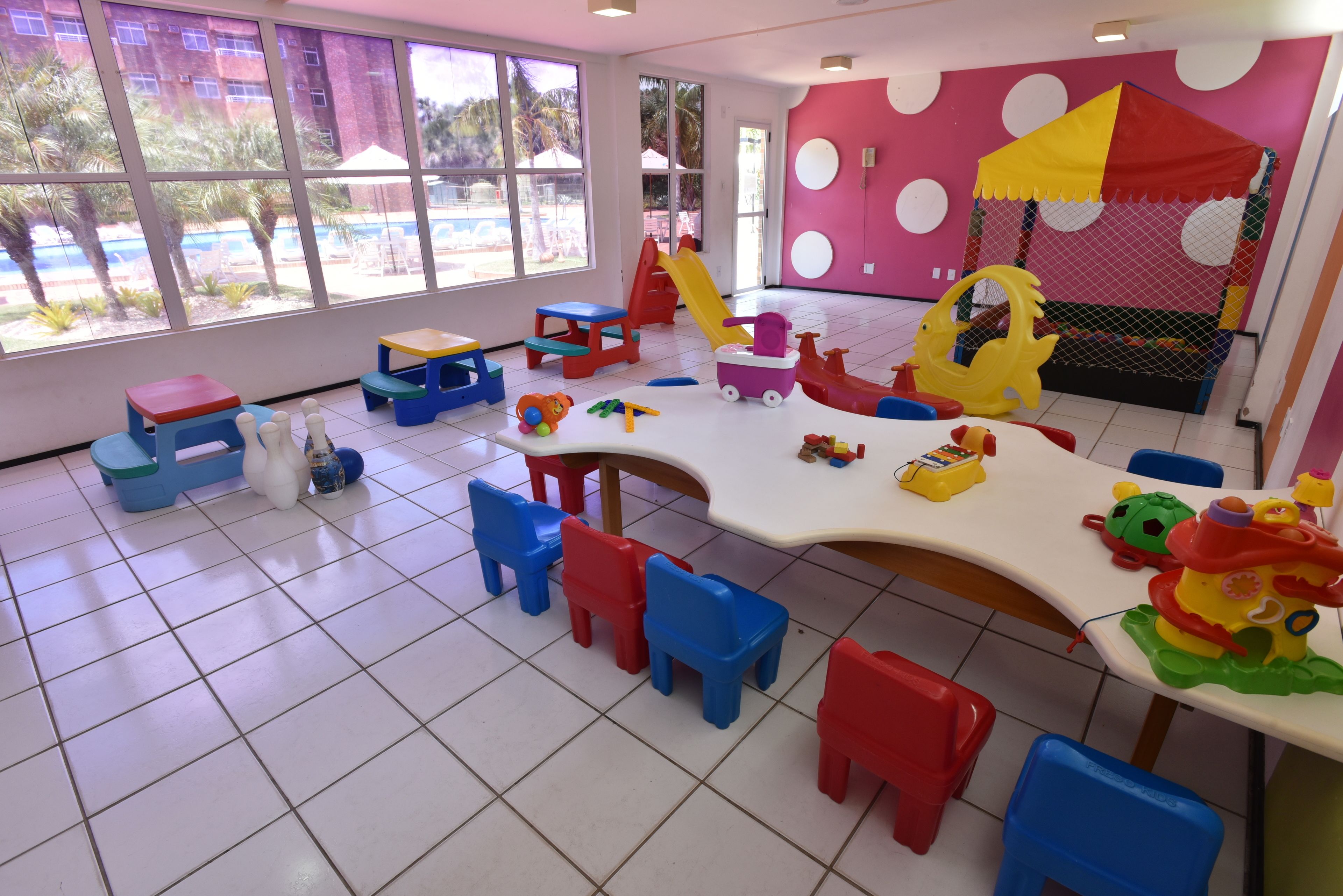 Un área de juegos interior bien iluminada, con un ventanal grande, una carpa de juegos y diversos juguetes y mobiliario.