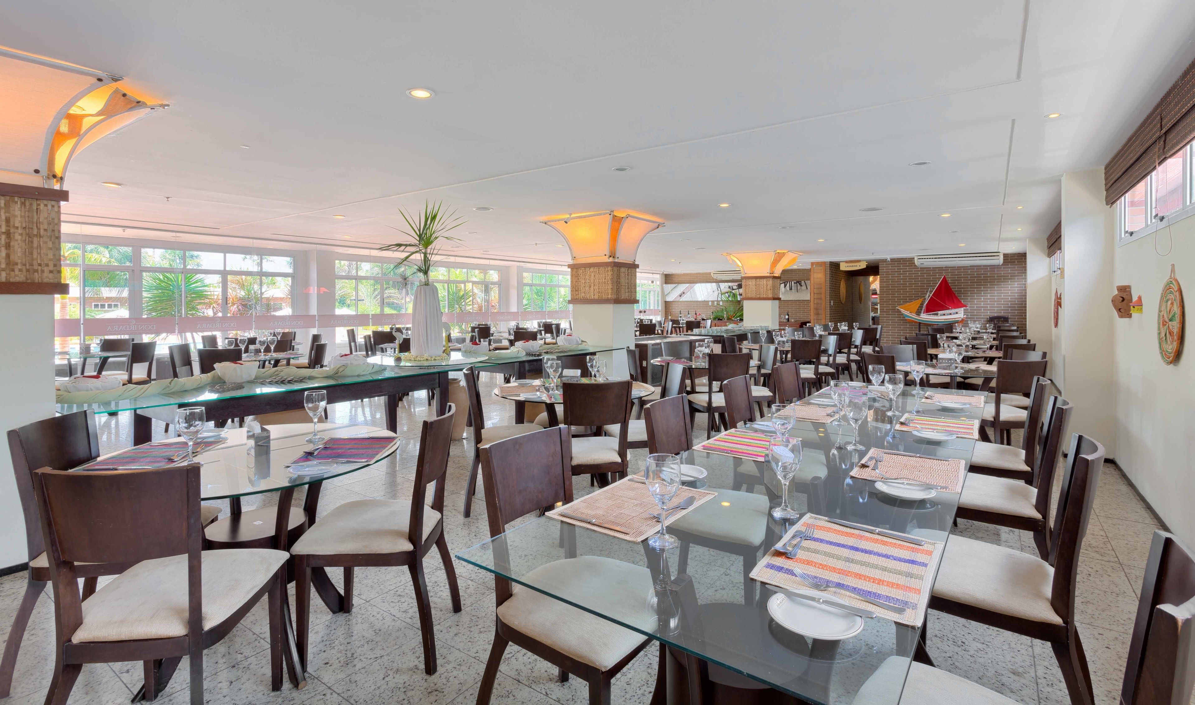 Un comedor amplio con varias mesas y sillas, ventanas grandes y una planta decorativa en el centro.