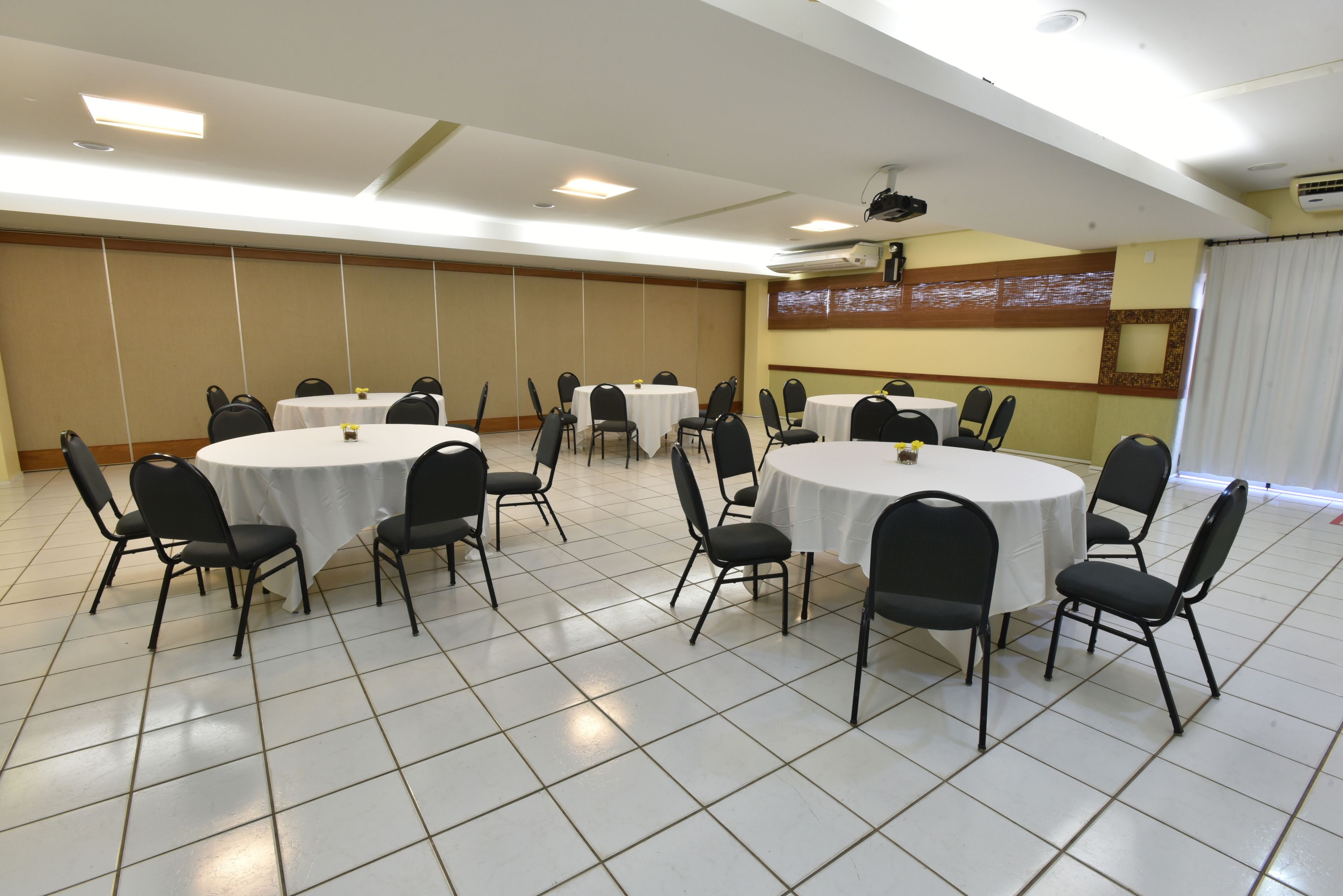 Une salle de conférence dotée de plusieurs tables rondes et de chaises noires, avec des murs blancs et un sol carrelé.