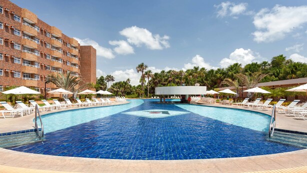 A swimming pool with lounge chairs and umbrellas, surrounded by palm trees and a hotel building. - Hotel Gran Lençóis Flat (Barreirinhas)