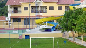 Um parquinho com um escorrega e uma quadra de basquete em frente a um edifício amarelo com telhado vermelho.