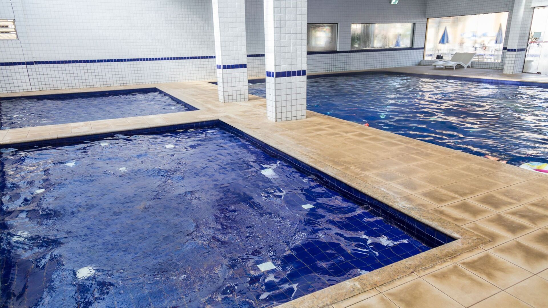 Una piscina interior con varias pistas de natación, paredes de azulejos y una zona de estar con sillas y sombrillas.