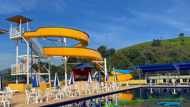 Um parque aquático com um grande escorregador amarelo, área de piscina com espreguiçadeiras e uma colina ao fundo.