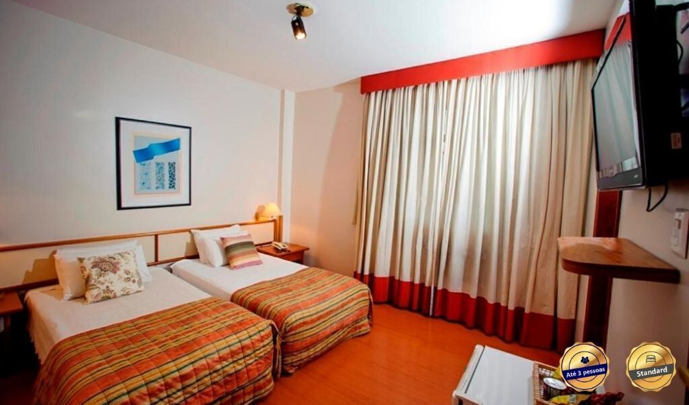 Quarto de hotel com duas camas, televisão, escrivaninha e janela com cortinas.