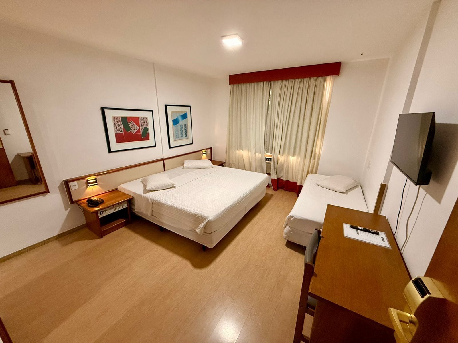 Quarto de hotel com uma cama grande, mesas de cabeceira, escrivaninha, uma cadeira, televisão e duas fotografias emolduradas na parede.