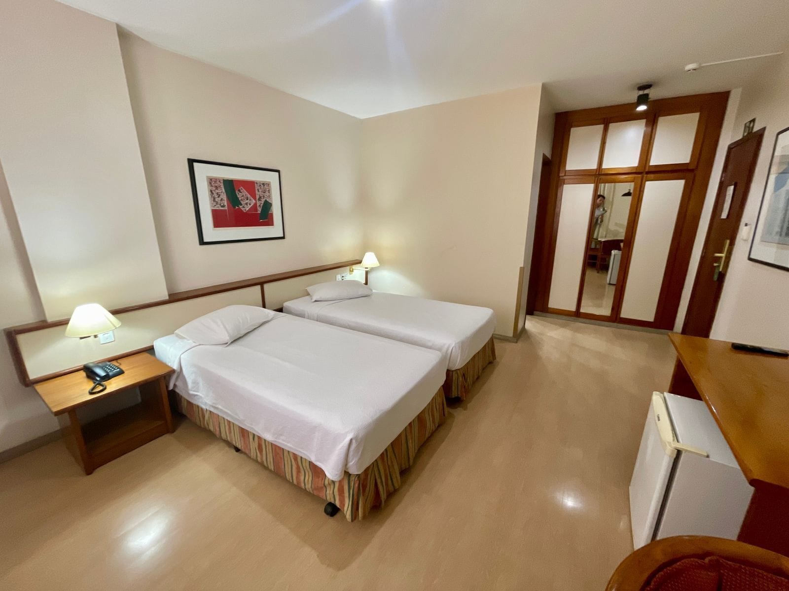A hotel room with two beds, a desk, a chair, a lamp, a phone, a mirror, and a door.