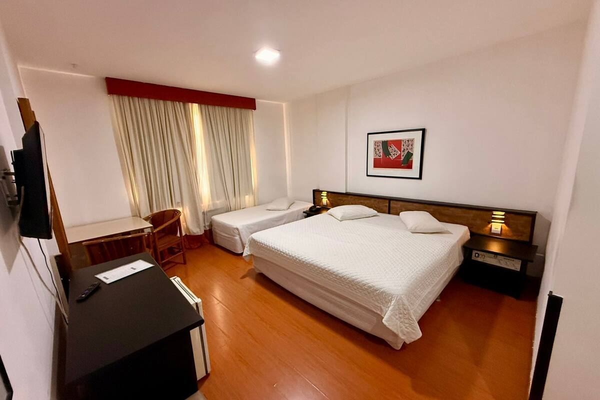 A hotel room with a desk, two single beds, a chair, a TV, and a painting on the wall.