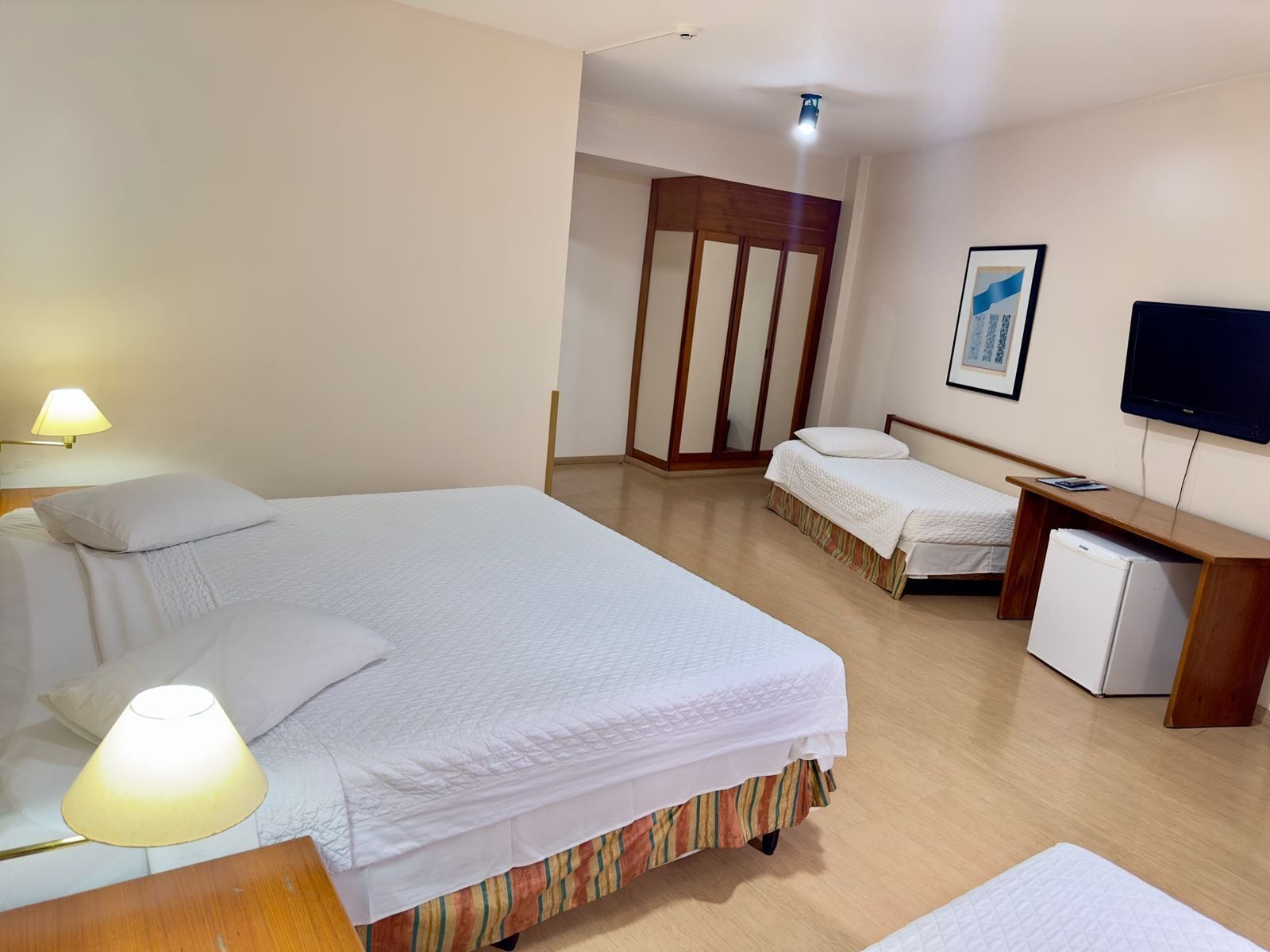 Habitación de hotel con una cama grande, una cama más pequeña, un escritorio con una computadora, un televisor y un refrigerador.