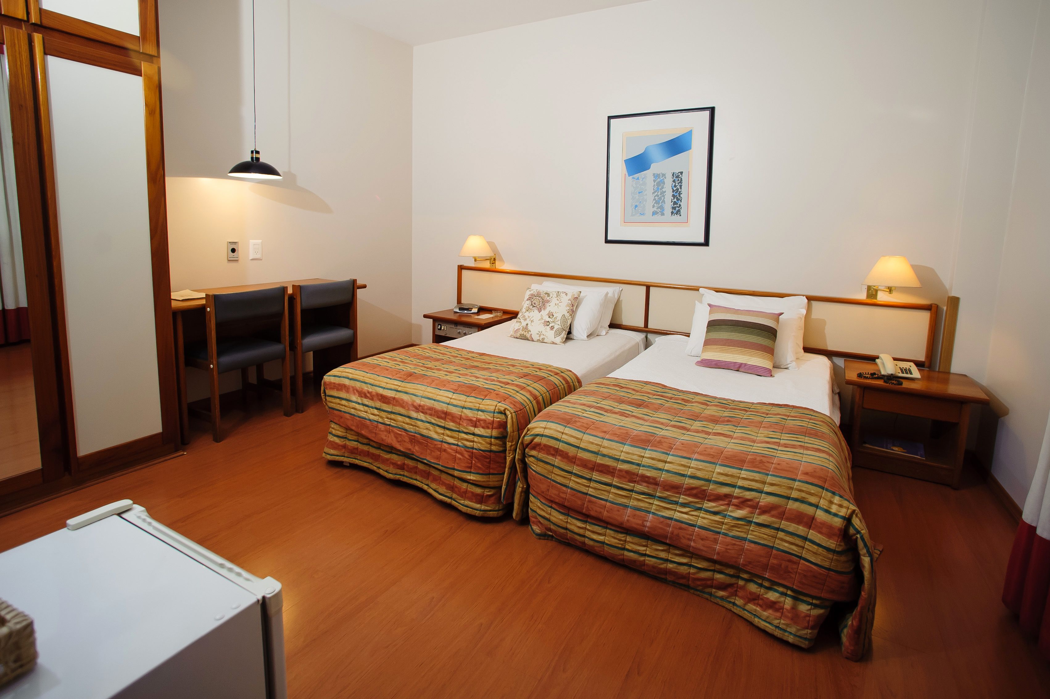 Quarto de hotel com duas camas, uma escrivaninha, uma cadeira, um abajur e um quadro na parede.