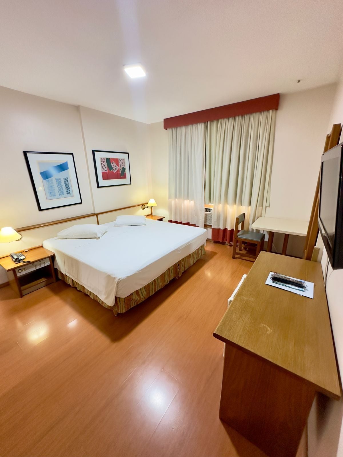 A hotel room with a large bed, a desk, a chair, a TV, and two framed pictures on the wall.