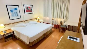 A hotel room with a large bed, a desk, a chair, a TV, and two framed pictures on the wall.