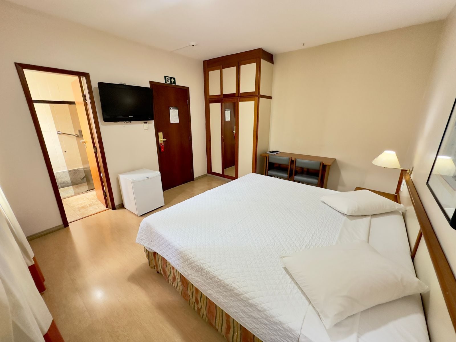 Quarto de hotel com uma cama, uma escrivaninha, uma cadeira, uma televisão e uma porta que dá para um banheiro.