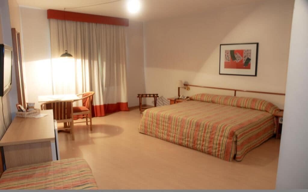 A hotel room with a bed, a desk, a chair, a bedside table, and a window with curtains.