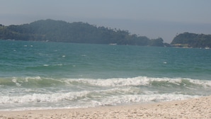 A beach with waves approaching the shore, a sandy beach, and a forested hill in the background. - Pousada The Goldeneye (Porto Belo)