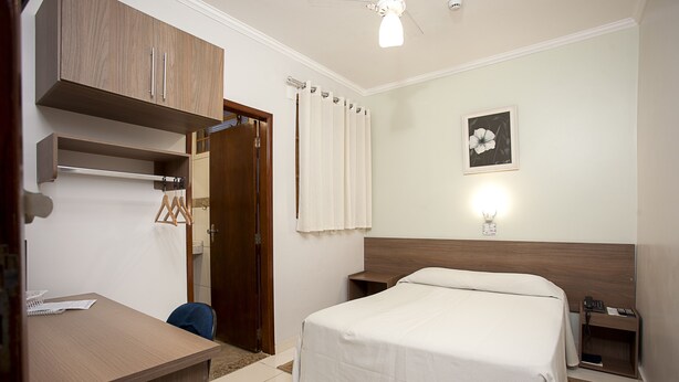 A neatly arranged hotel room with a bed, bedside table, and a desk with a chair. - Alves Hotel (Marilia)