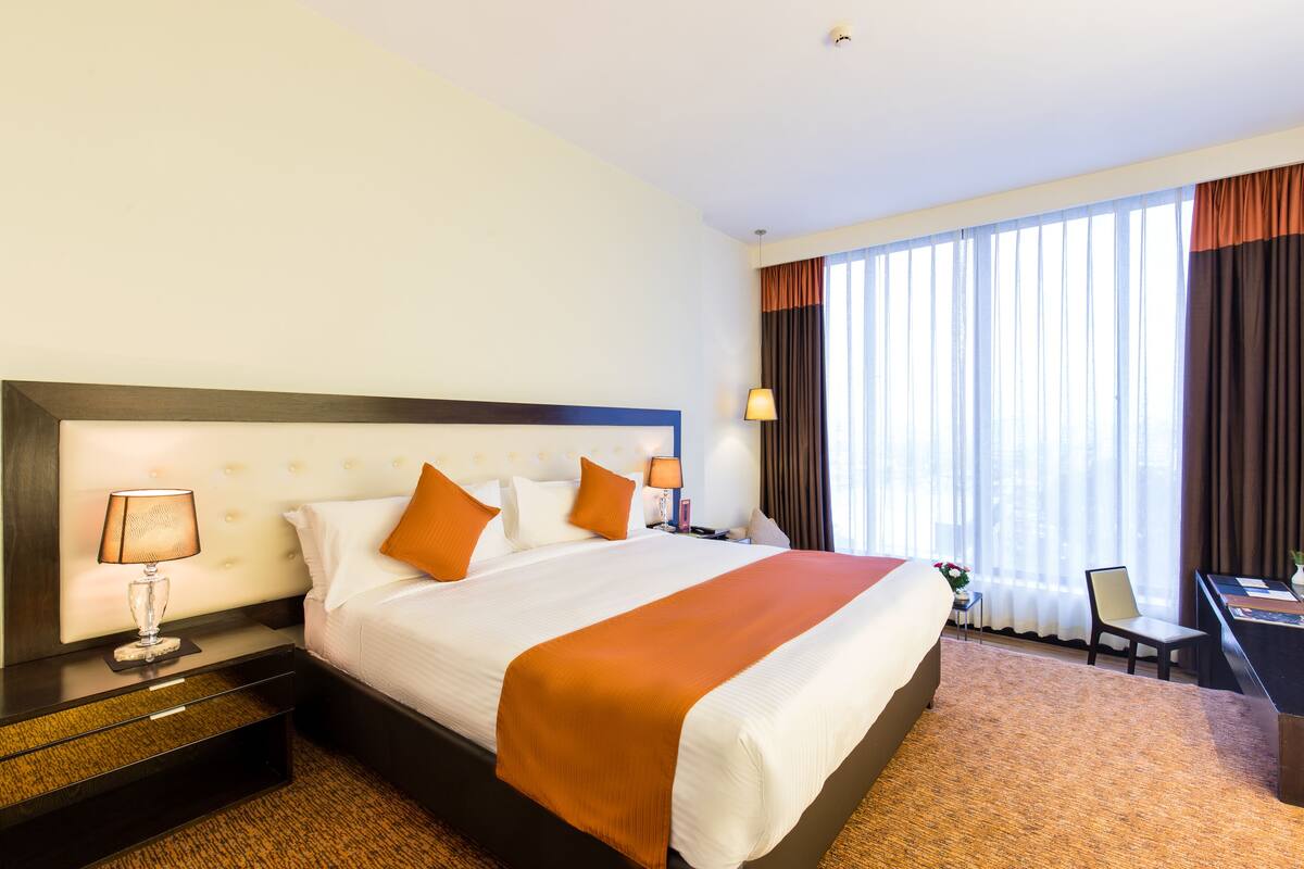 Habitación de hotel con una cama grande, una mesita de noche con lámpara, un escritorio con silla y una ventana con cortinas.