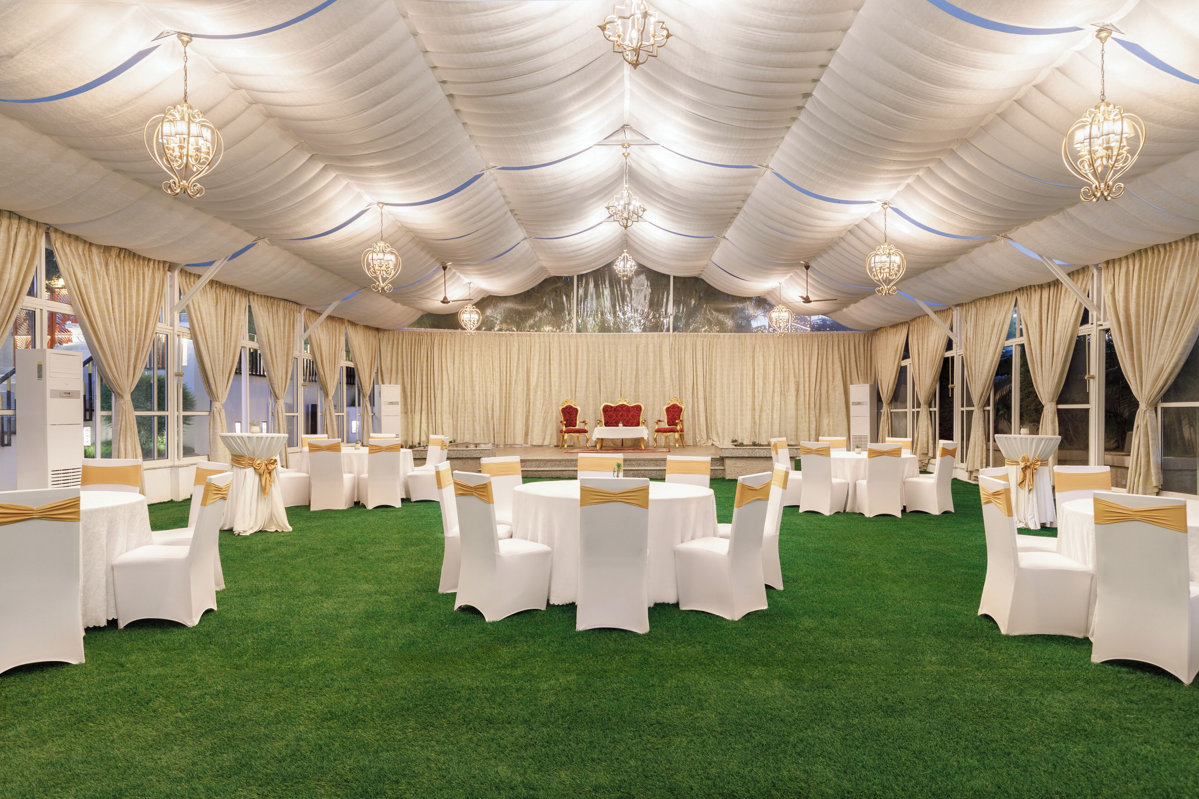 Un espacio para eventos con carpas, mesas redondas y sillas, un escenario con sillas rojas y candelabros.