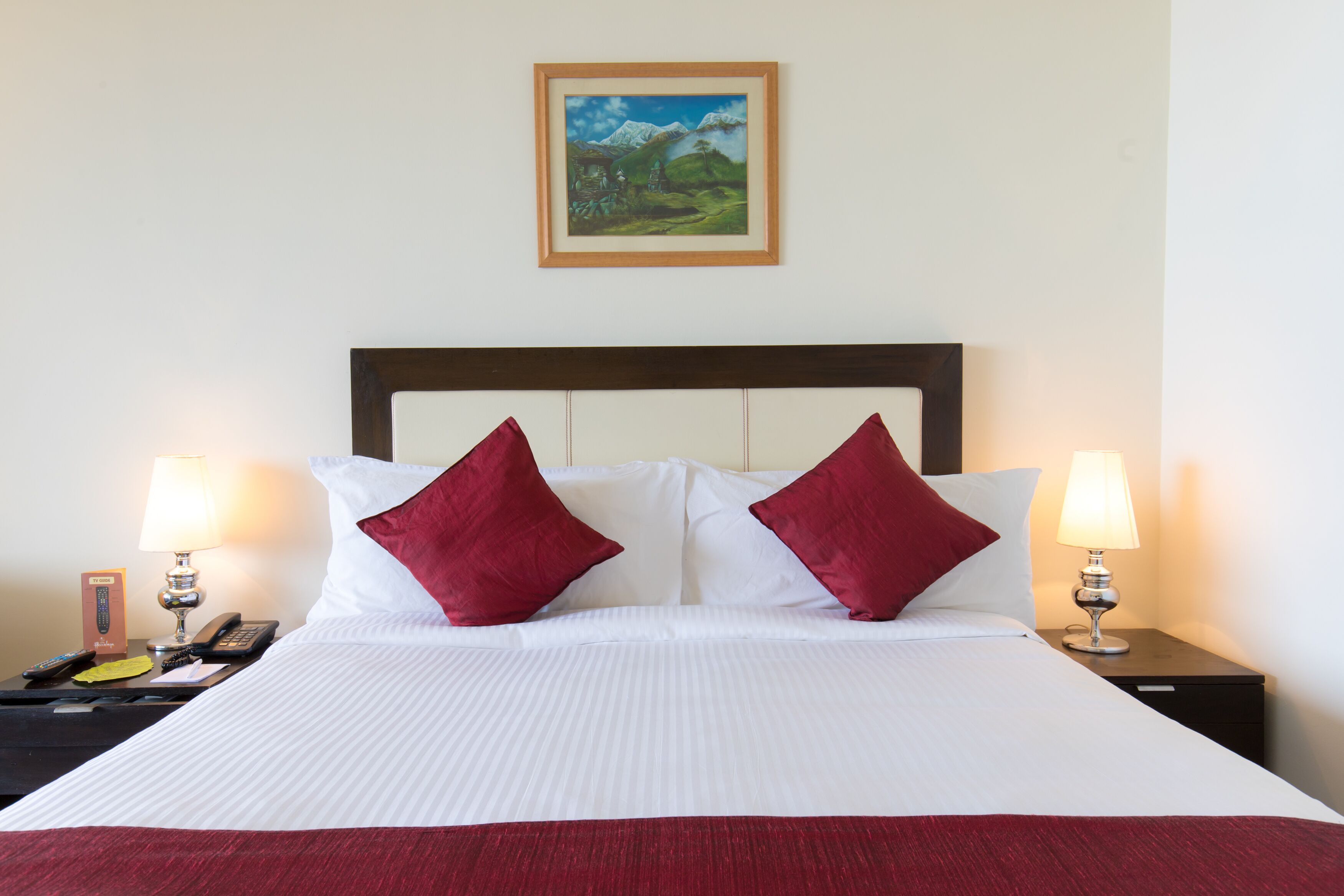 Una cama bien hecha con sábanas blancas y dos almohadas rojas, flanqueada por mesitas de noche con lámparas y un control remoto.