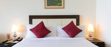 Una cama bien hecha con sábanas blancas y dos almohadas rojas, flanqueada por mesitas de noche con lámparas y un control remoto.