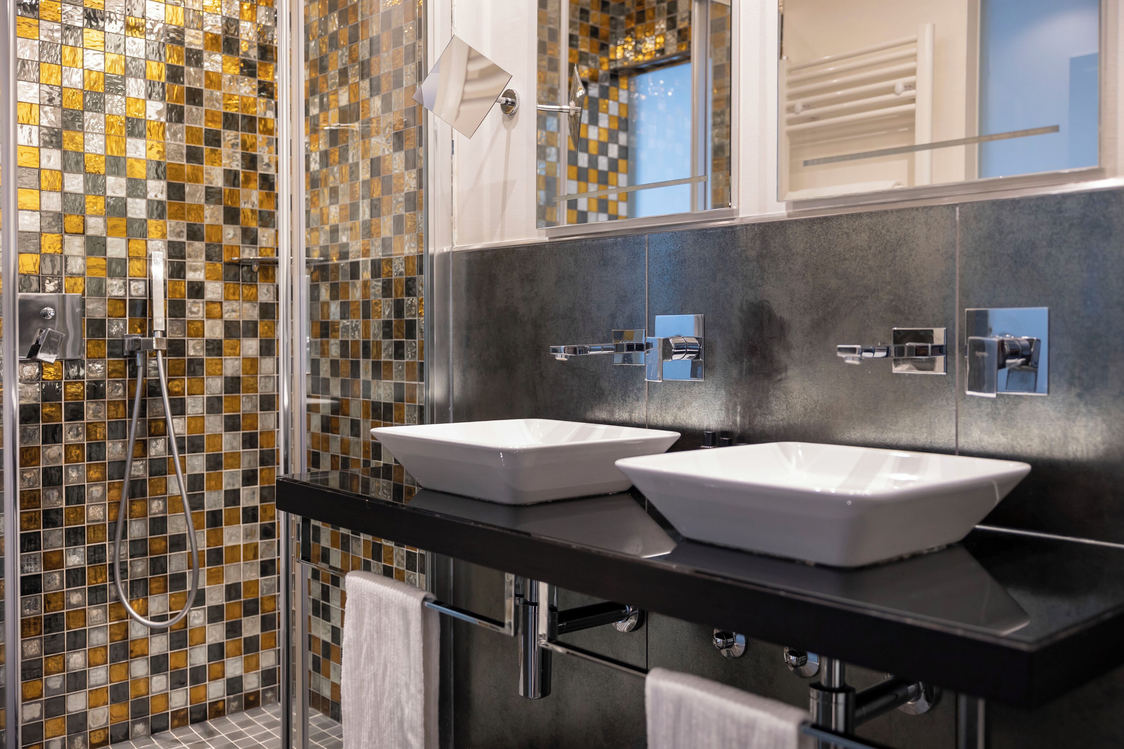 A modern bathroom with two white sinks, a glass shower enclosure, and mosaic tile walls.
