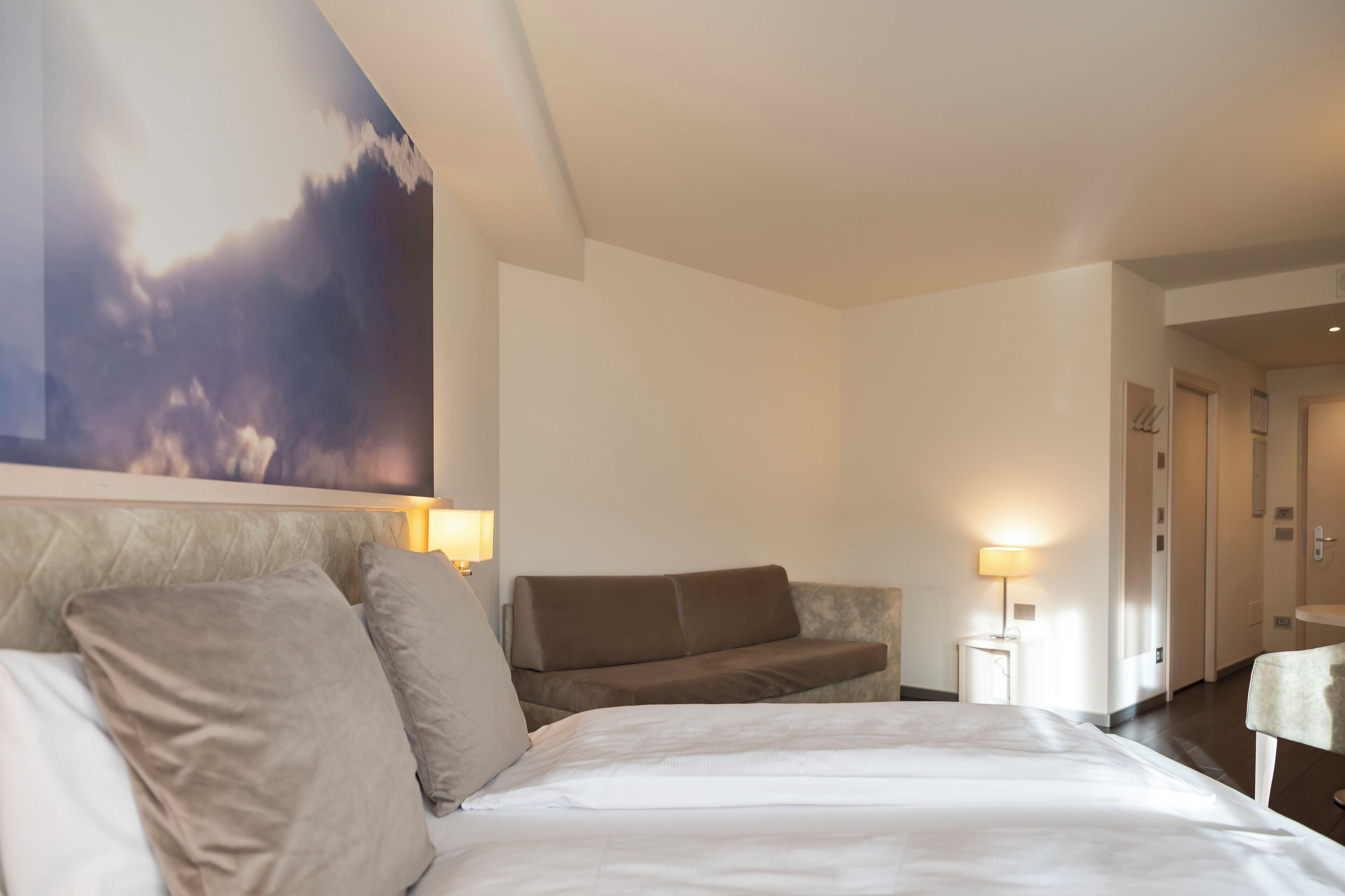 A hotel room with a bed, a sofa, a wall-mounted TV, and a bedside table with a lamp.