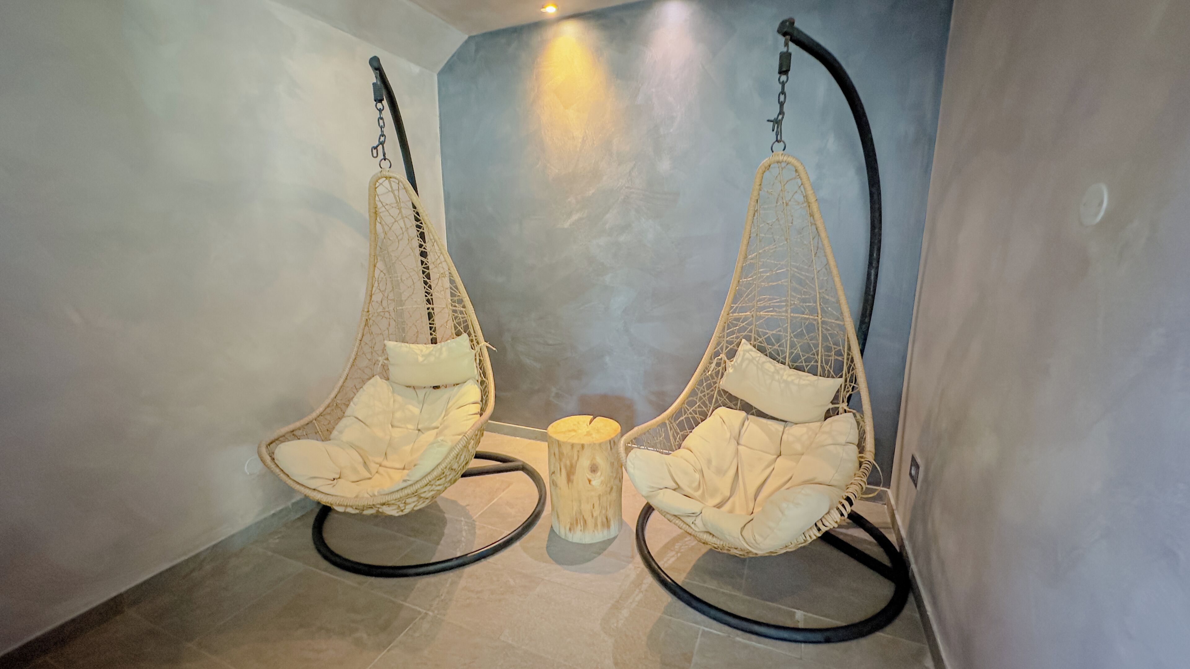Two hanging chairs with cushions, a wooden stool, and a wall-mounted light fixture in a room with tiled flooring.