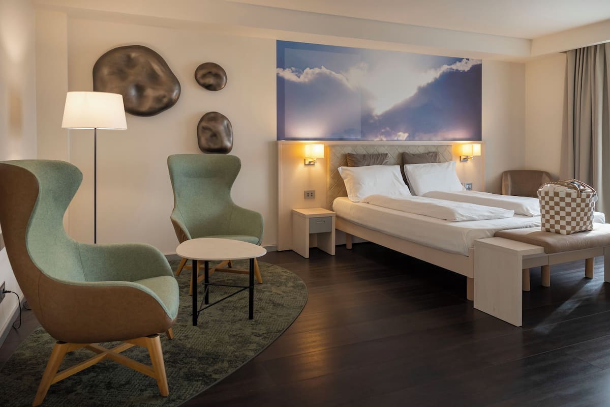 A modern hotel room with a bed, two armchairs, a floor lamp, and a painting of clouds.