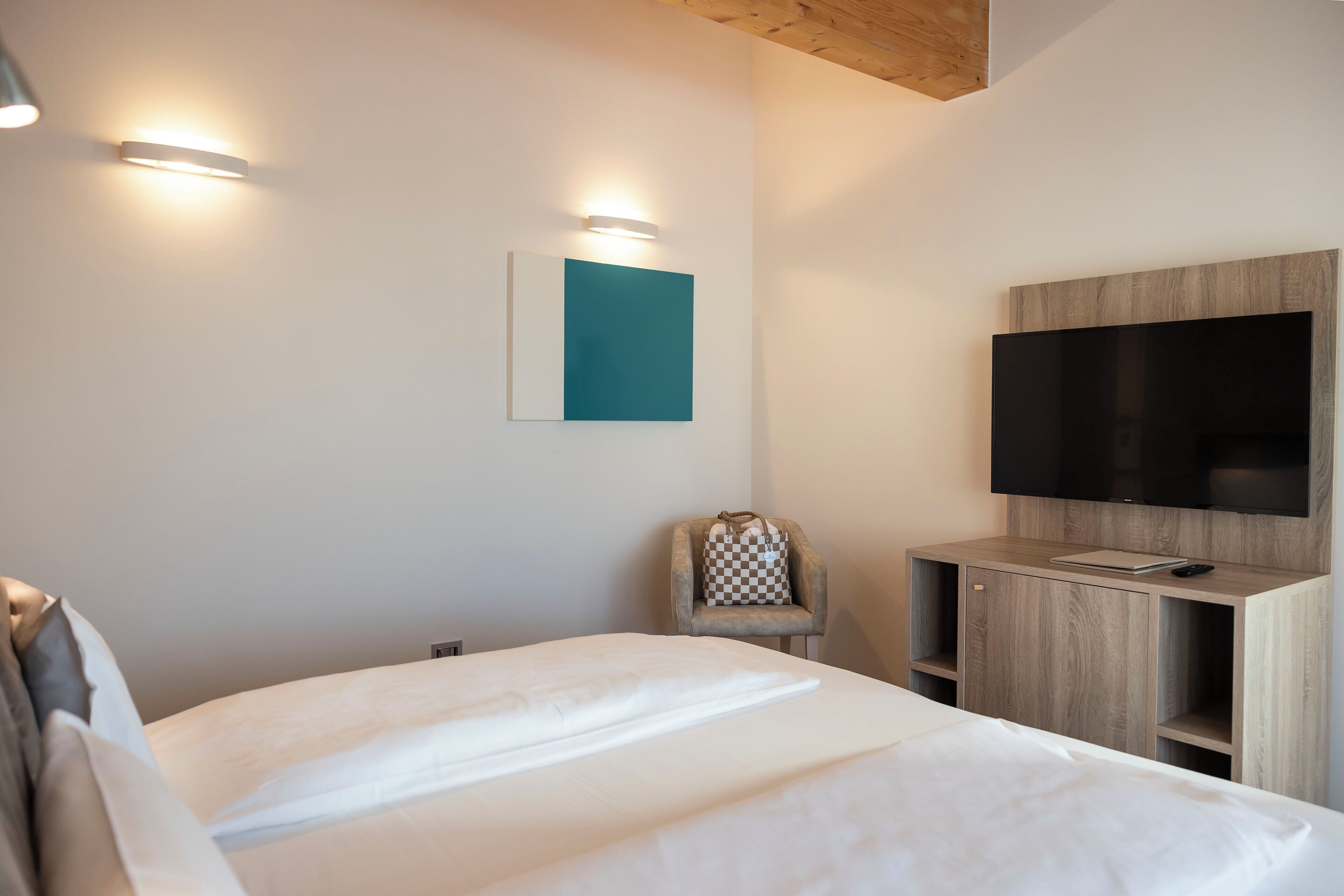 Una moderna camera d'albergo con un letto grande, una TV a schermo piatto montata su un mobile in legno e una poltrona con cuscino a quadretti.