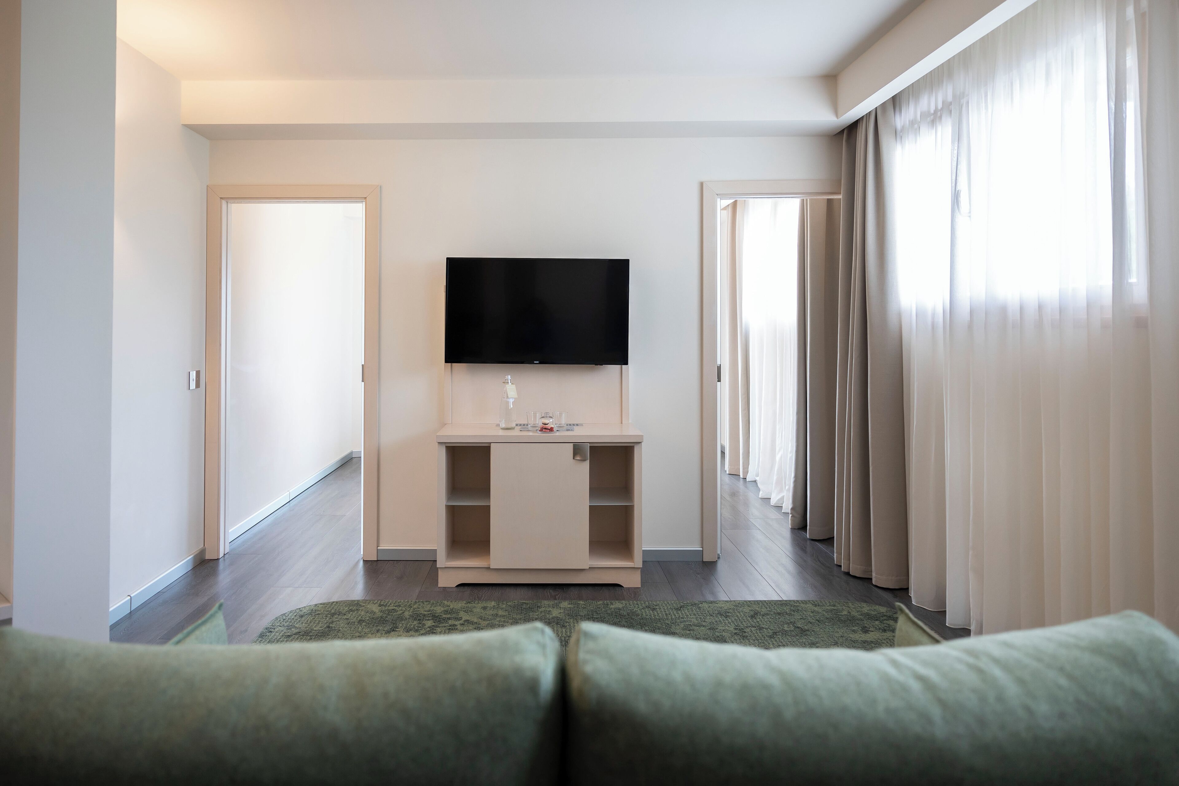 Una habitación de hotel moderna con un televisor de pantalla plana montado sobre una consola de color claro, un tapete verde y un sofá verde.