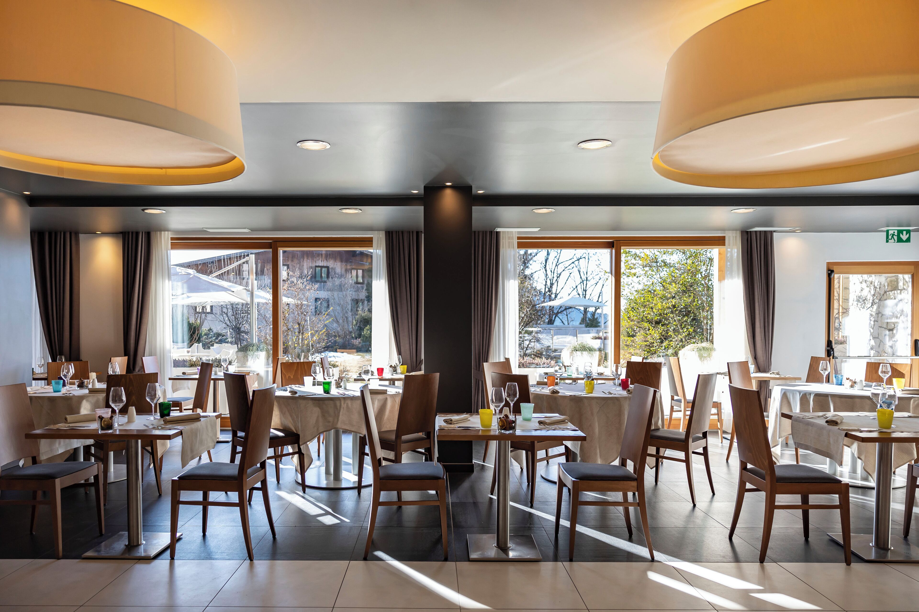 Een moderne eetruimte met meerdere tafels en stoelen, grote ramen voorzien van gordijnen en uitzicht op buiten.