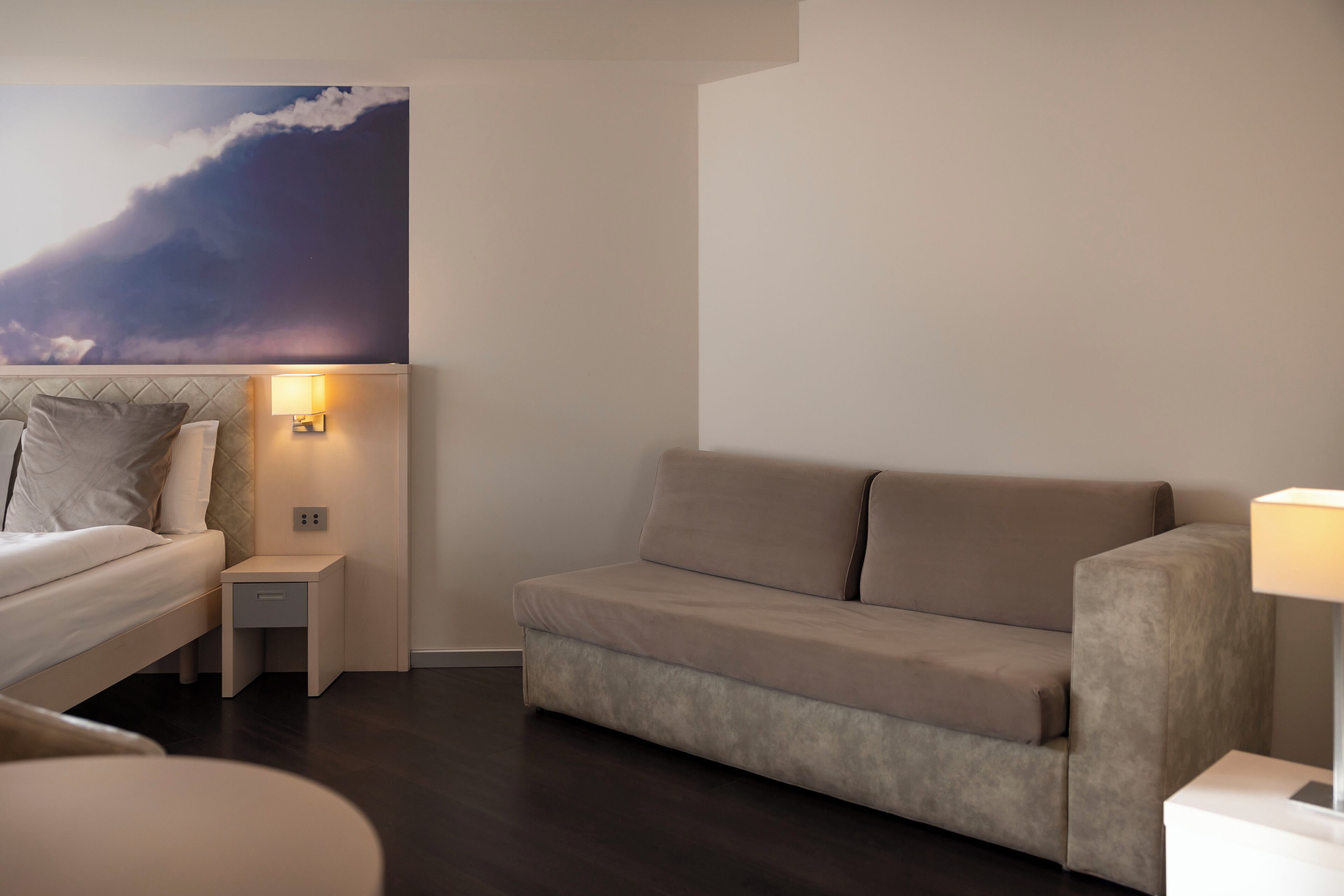 A modern hotel room with a bed, a sofa, a nightstand with a lamp, and a wall mural.