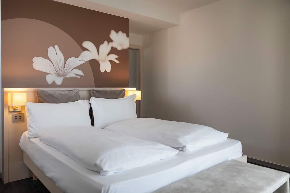 A neatly made bed with white linens, a wall-mounted light fixture, and a large floral artwork above the bed.