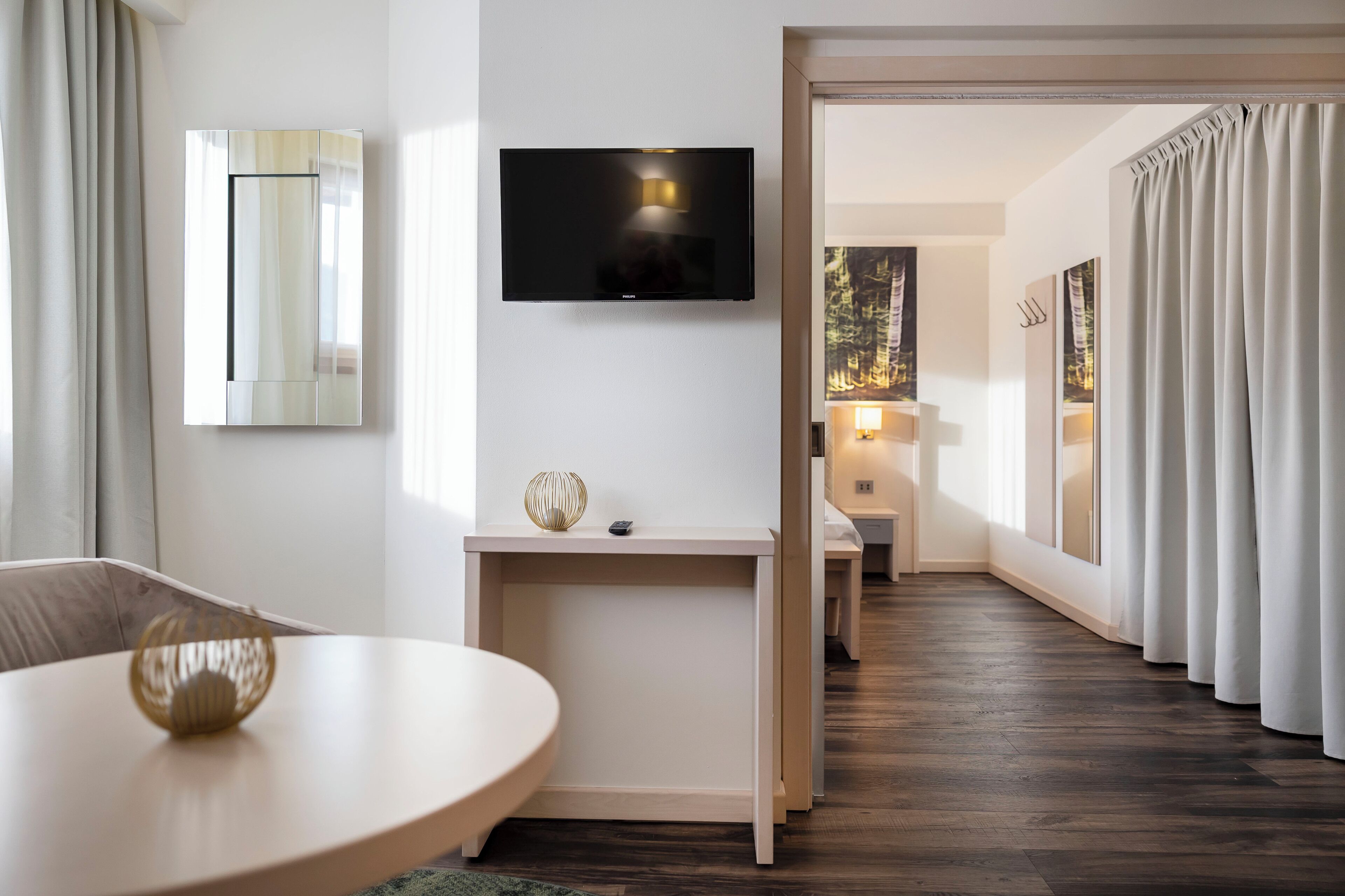 A modern hotel room with a flat-screen TV mounted on the wall, a wooden desk with a lamp, and a view of a forest through a window.