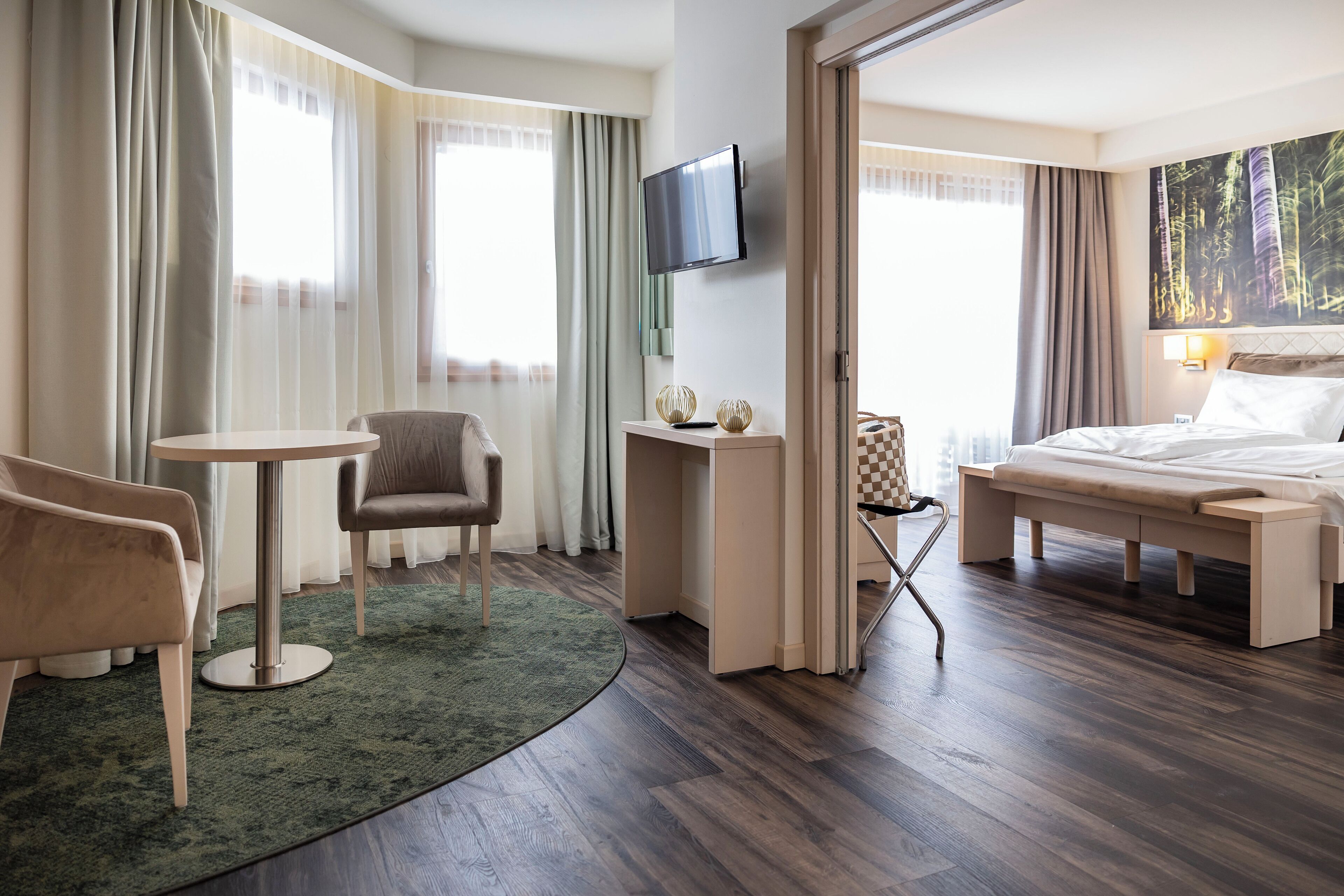 Quarto de hotel com cama, escrivaninha, cadeira, televisão e uma janela grande com cortinas.