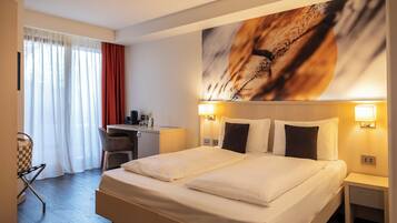 A hotel room with a double bed, a desk with a chair, a small table, and a large window with curtains.