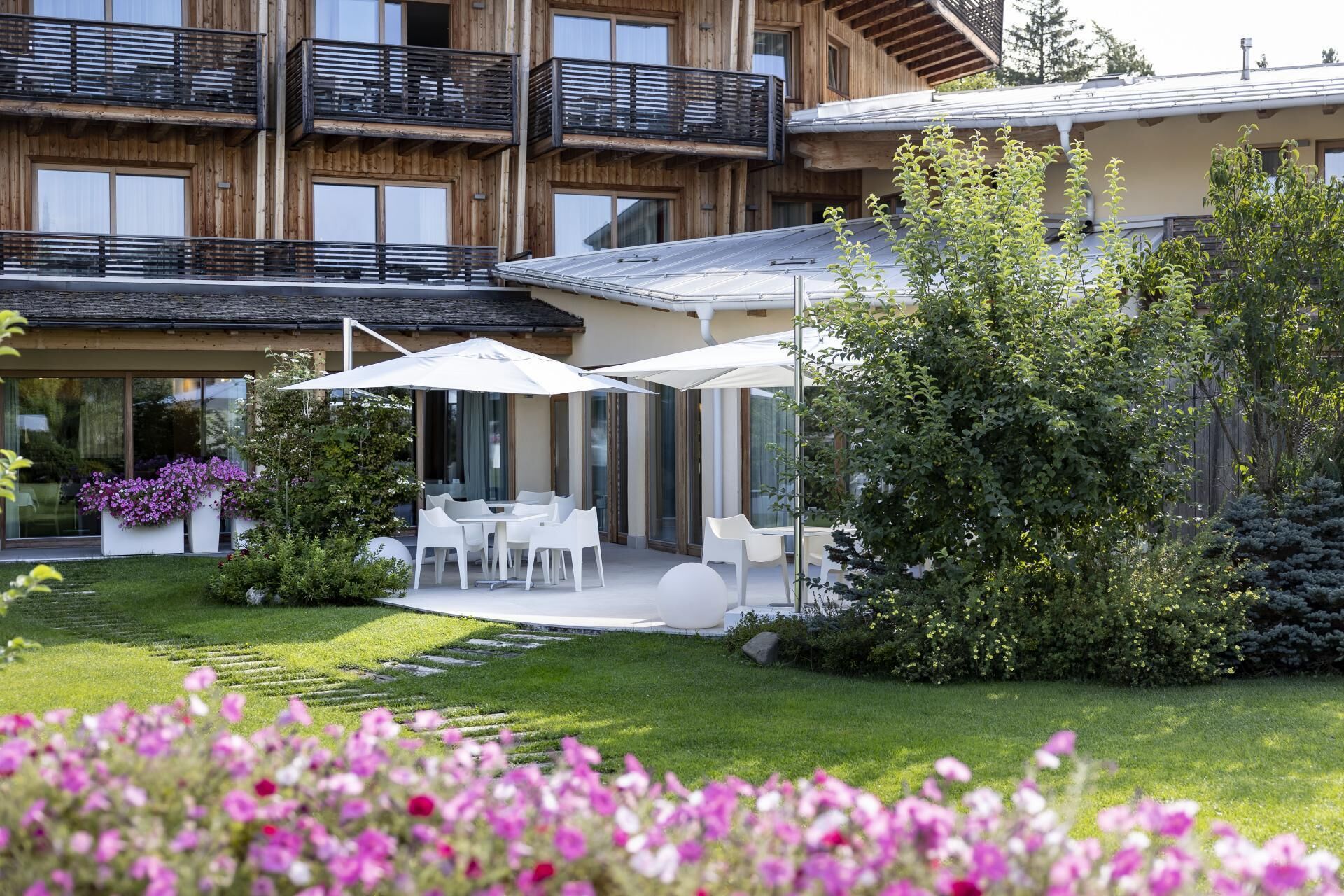 Un edificio moderno con fachada de madera, zona de estar al aire libre con sillas y sombrillas blancas, y un jardín cuidado con diversas plantas y flores.