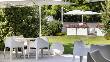 Un espace extérieur aménagé en terrasse, avec des tables et des chaises blanches, entouré de verdure et agrémenté de parasols.