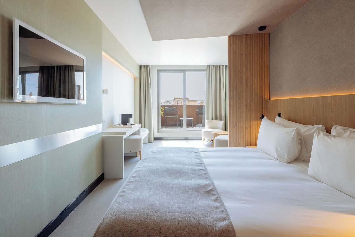 Hotellihuone, jossa on suuri sänky, työpöytä, tuoli ja ikkunasta avautuva näkymä kaupunkiin.