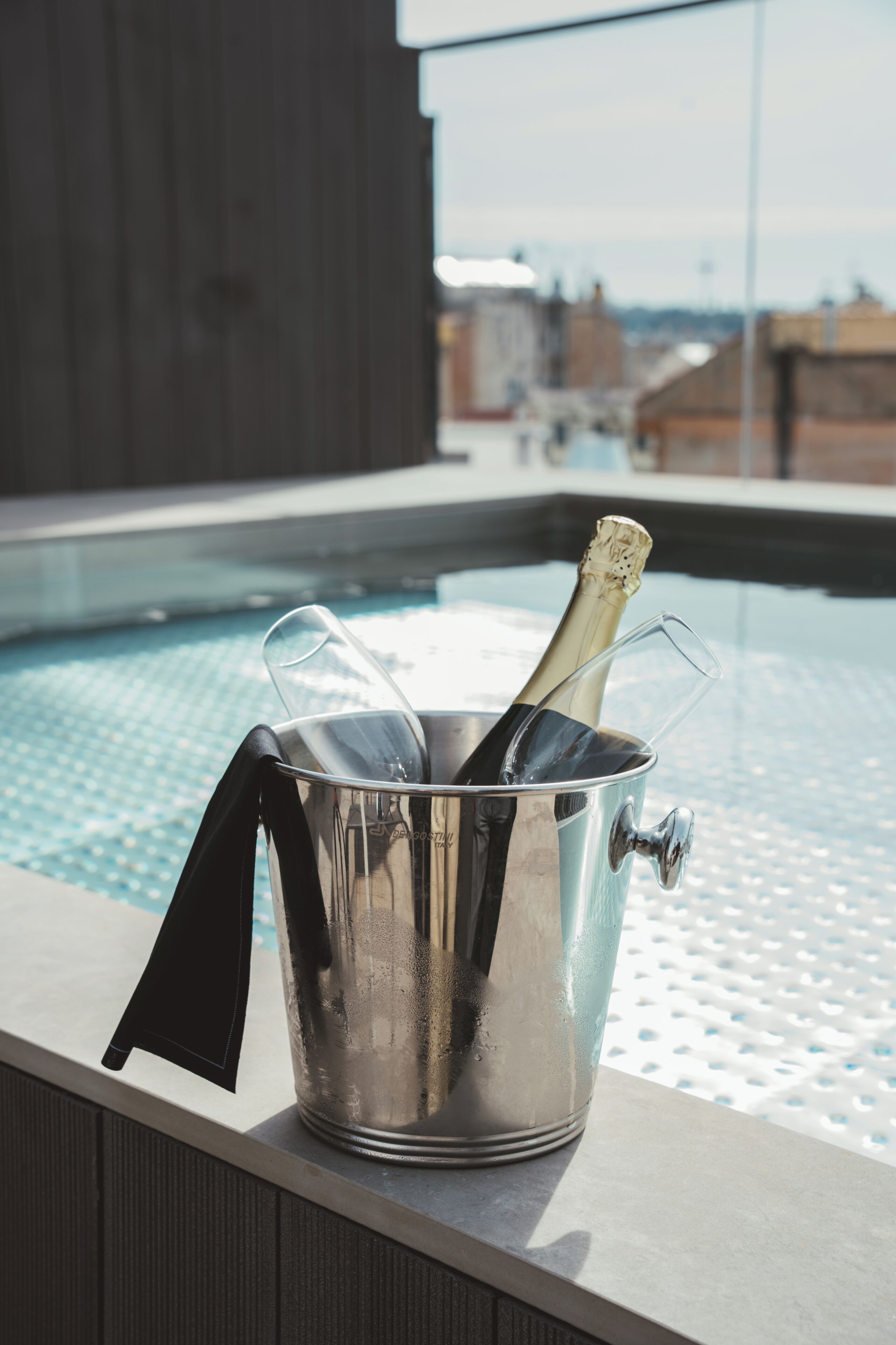 A metallic bucket with a bottle and glass on a balcony overlooking a swimming pool.