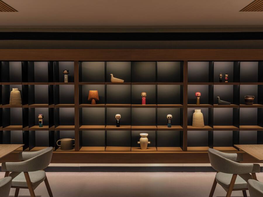 A modern interior with a long, dark wooden bookshelf filled with various decorative items. There are two wooden tables with chairs in front.