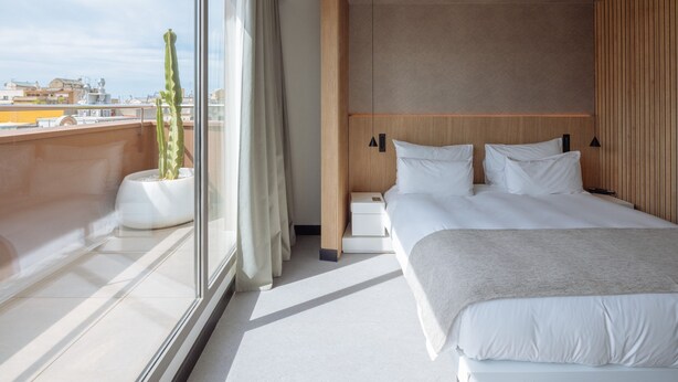 Moderni hotellihuone, jossa on suuri sänky, yöpöydät, ruukkukasvi ja suuret ikkunat, joista avautuu näkymä kaupunkiin.