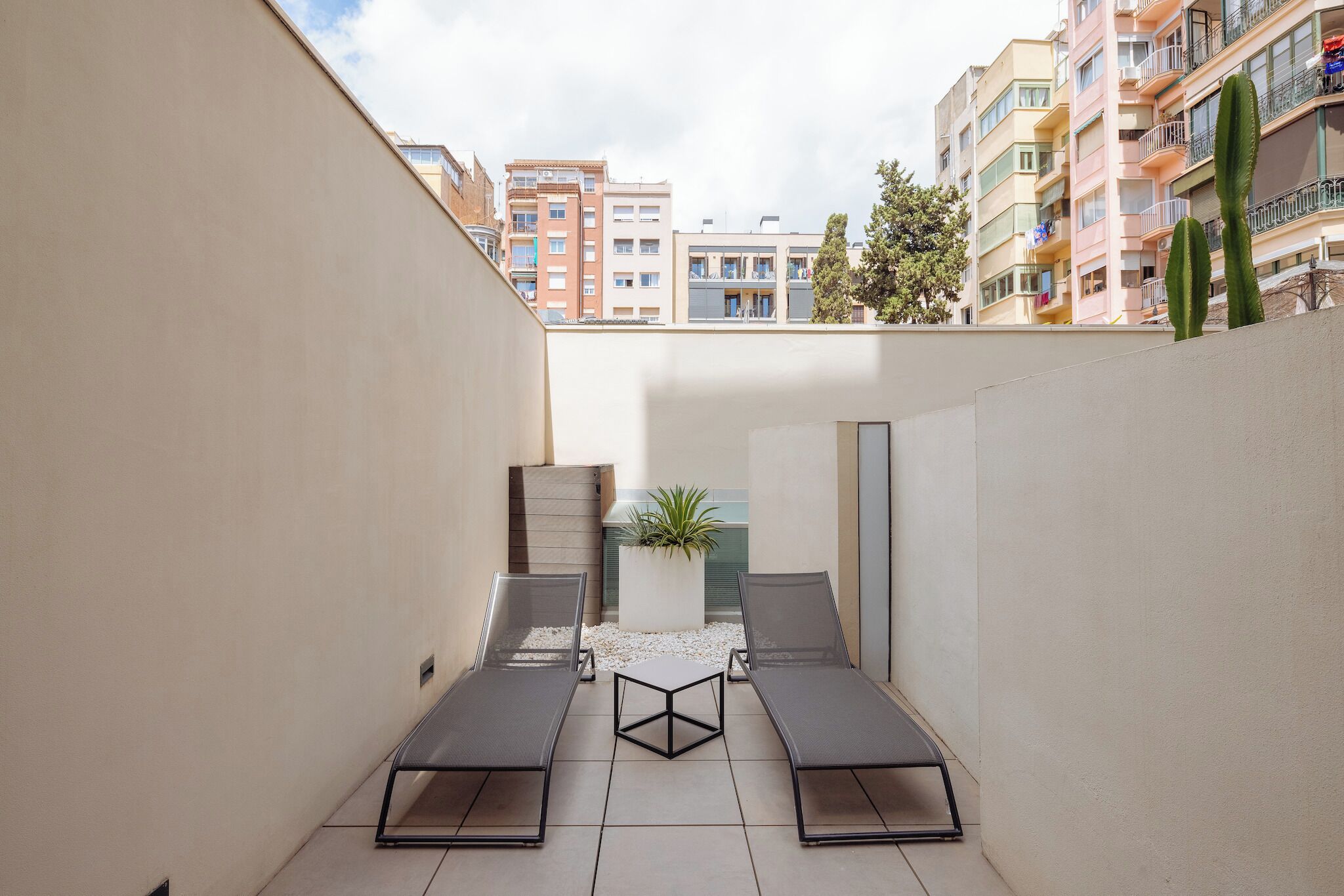 A small outdoor patio with two lounge chairs and a table, surrounded by a low wall and a potted plant.