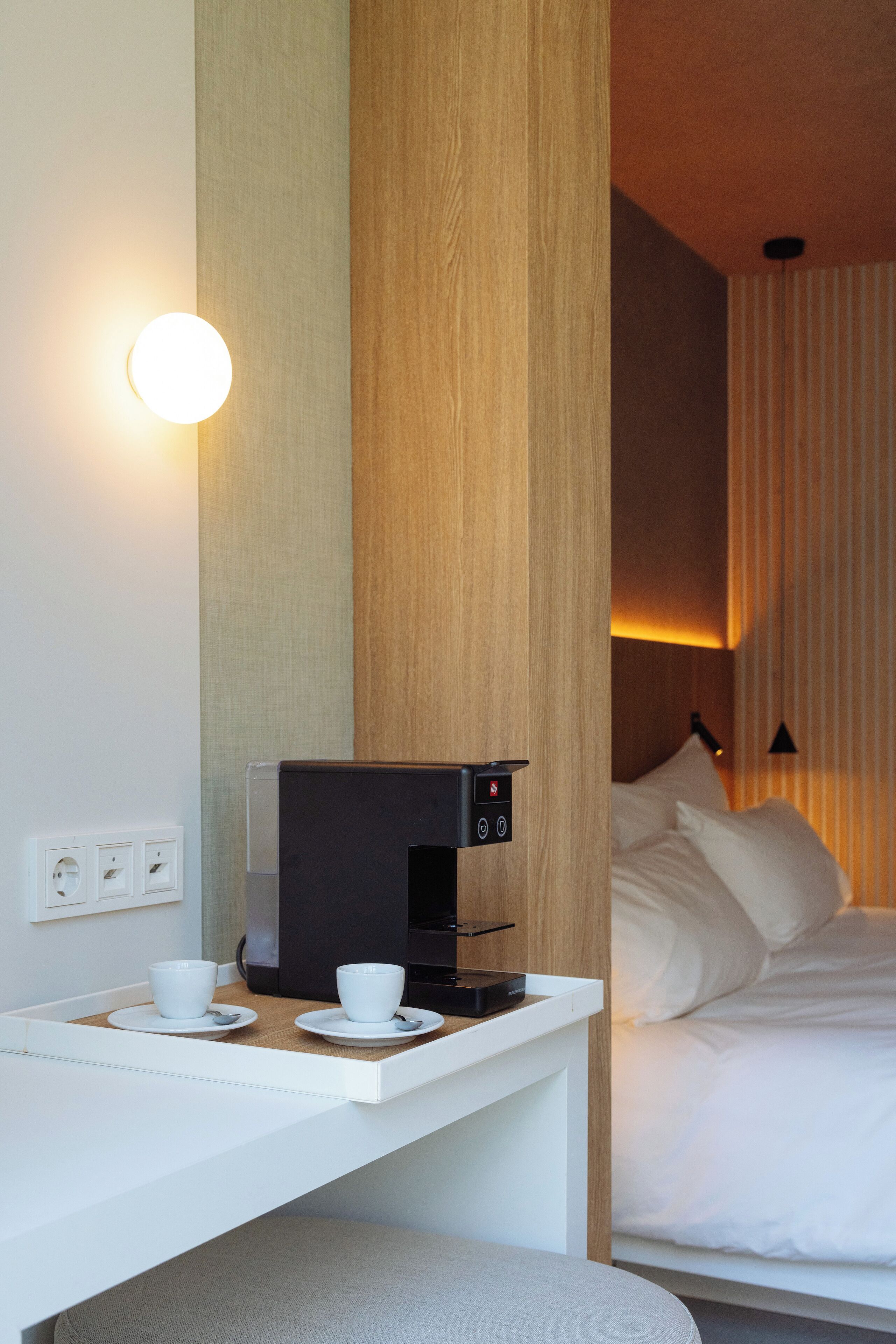 A hotel room with a bed, a bedside table with a coffee machine, and a wall-mounted light.
