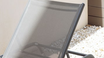 Una moderna silla de lounge para exterior con respaldo de malla y estructura metálica, ubicada en un patio con guijarros blancos y una pared de madera al fondo.