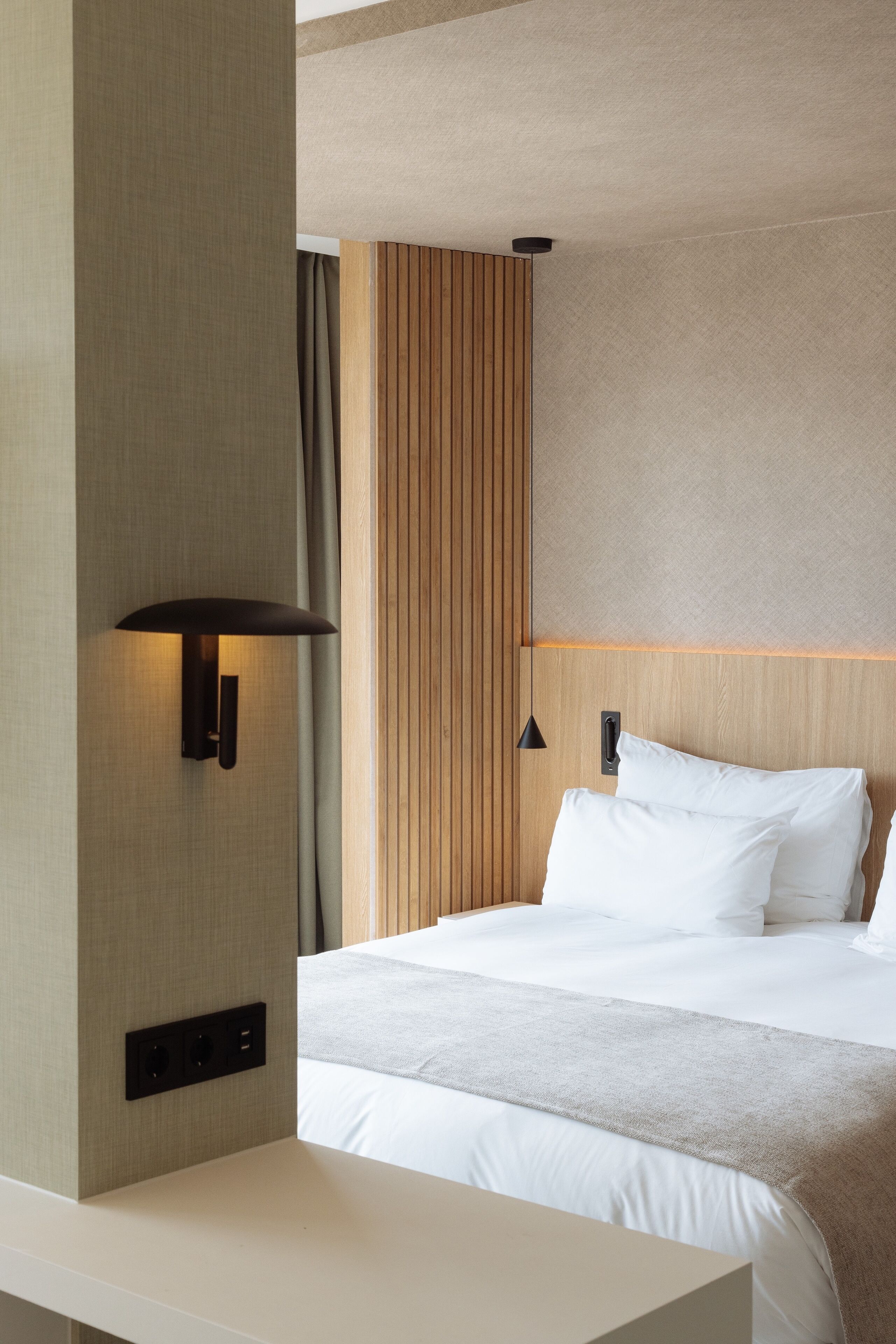 Moderni hotellihuone, jossa on suuri sänky, yöpöytä, seinään kiinnitetty valaisin ja puupaneelilla viimeistelty seinä.