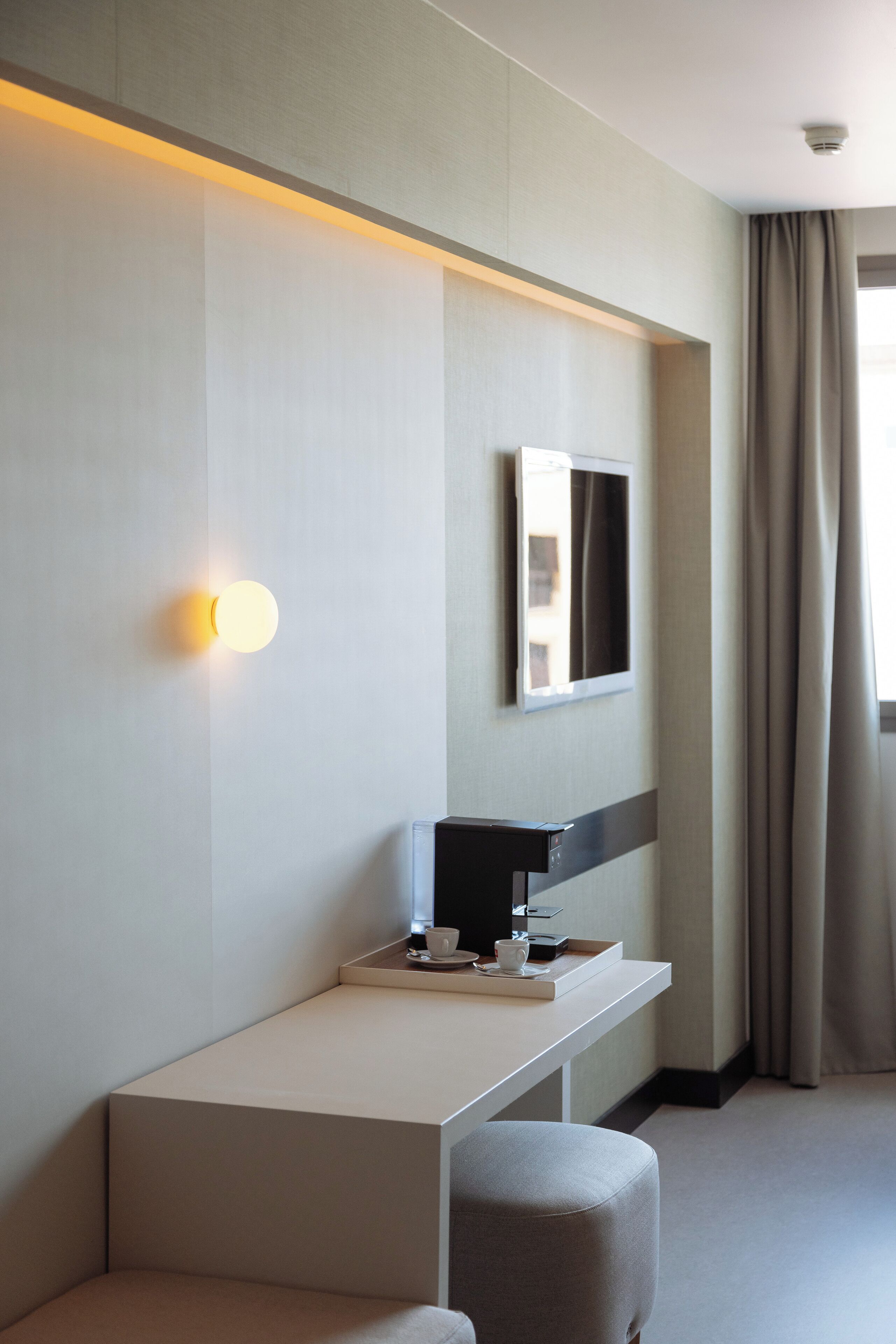 A modern hotel room with a minimalist design, featuring a flat-screen TV mounted on the wall, a small desk with a cup and a tray, and a comfortable seating area.