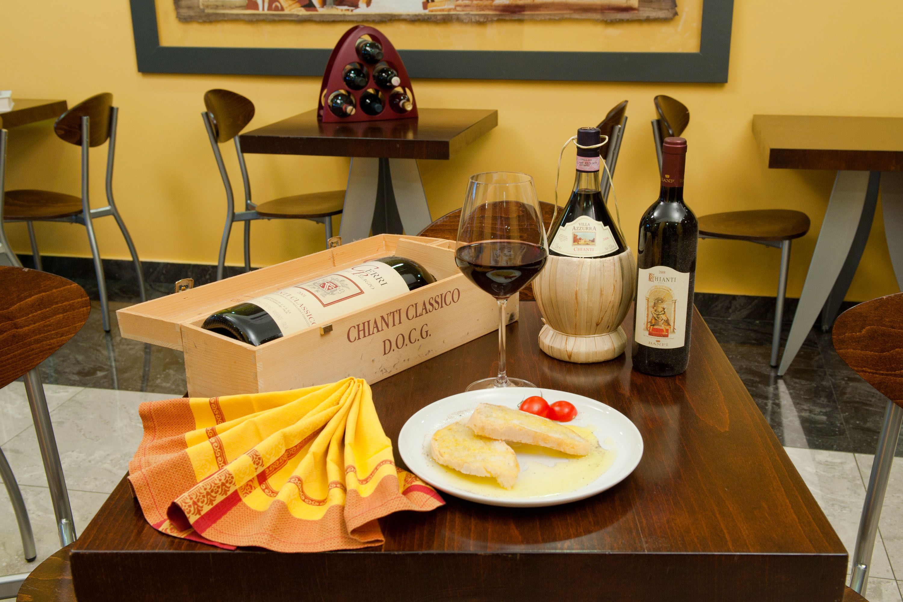 A dining table with a box of Chianti Classico wine, a bottle of Chianti, a glass of red wine, a plate with fish and cherry tomatoes, and a yellow cloth.