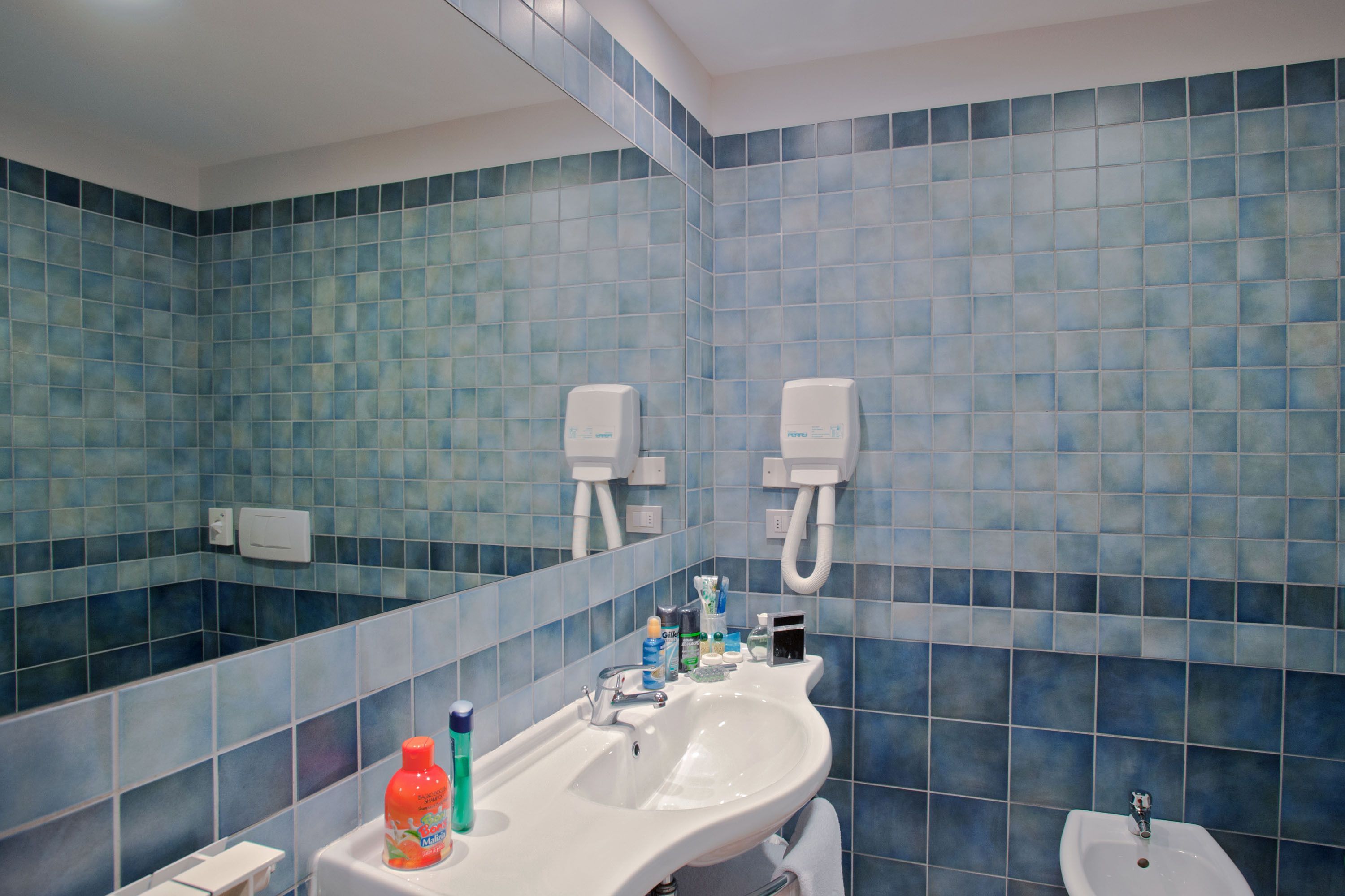 Baño con azulejos azules, un lavamanos, un espejo y dos secamanos.